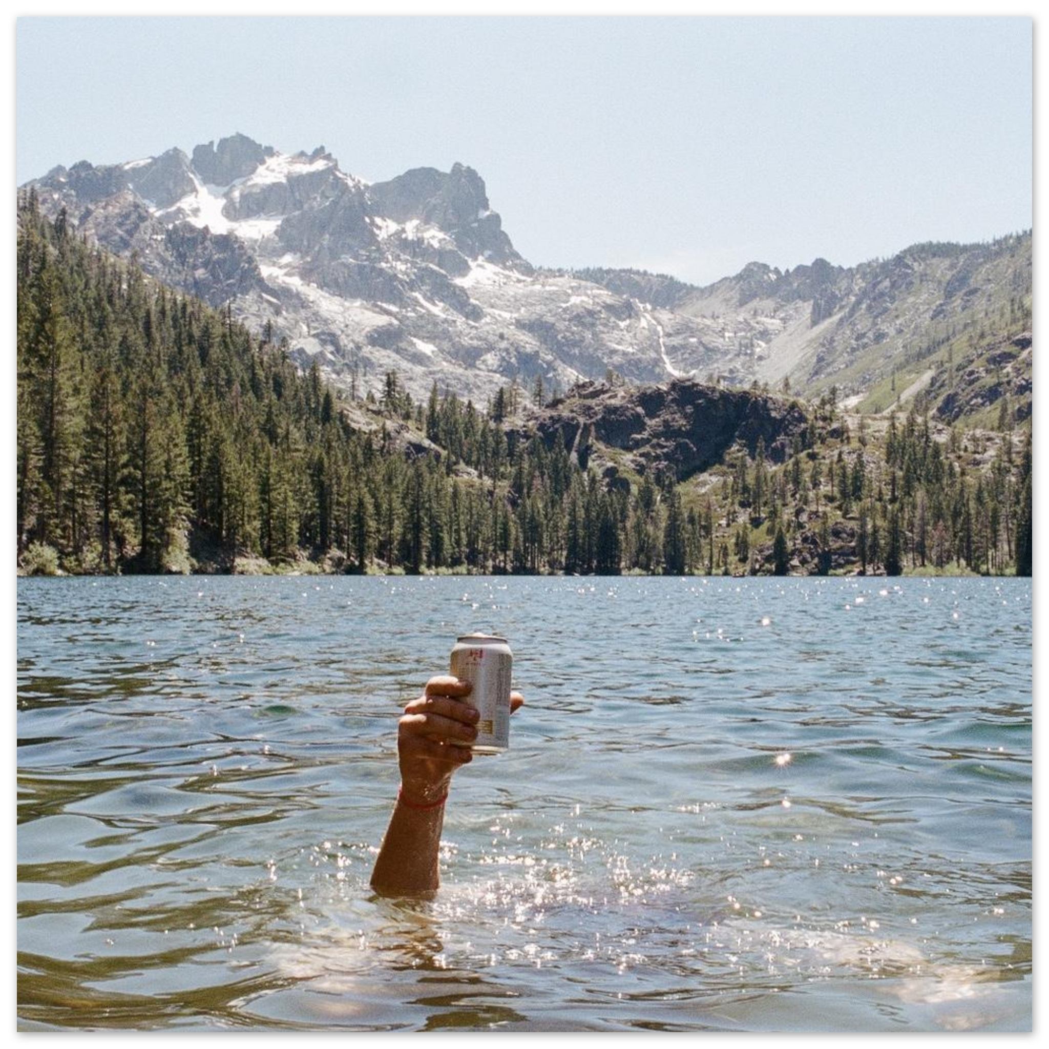Adventure photography print of hand holding beer in alpine lake with mountains and trees, lifestyle wall art